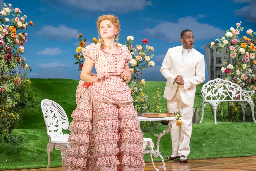 A woman in a pink floral Victorian dress with ruffled layers (portraying Gwendolen) stands in the foreground holding a letter, looking thoughtful. Behind her, a man in a white suit (played by Ncuti Gatwa as Algernon) stands near a white garden bench, looking toward her with a surprised expression.